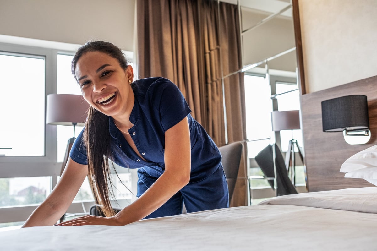 A female hotel staff member is preparing a room for the next guests. She carefully makes the bed, arranges pillows, and ensures the room is tidy and ready for service.